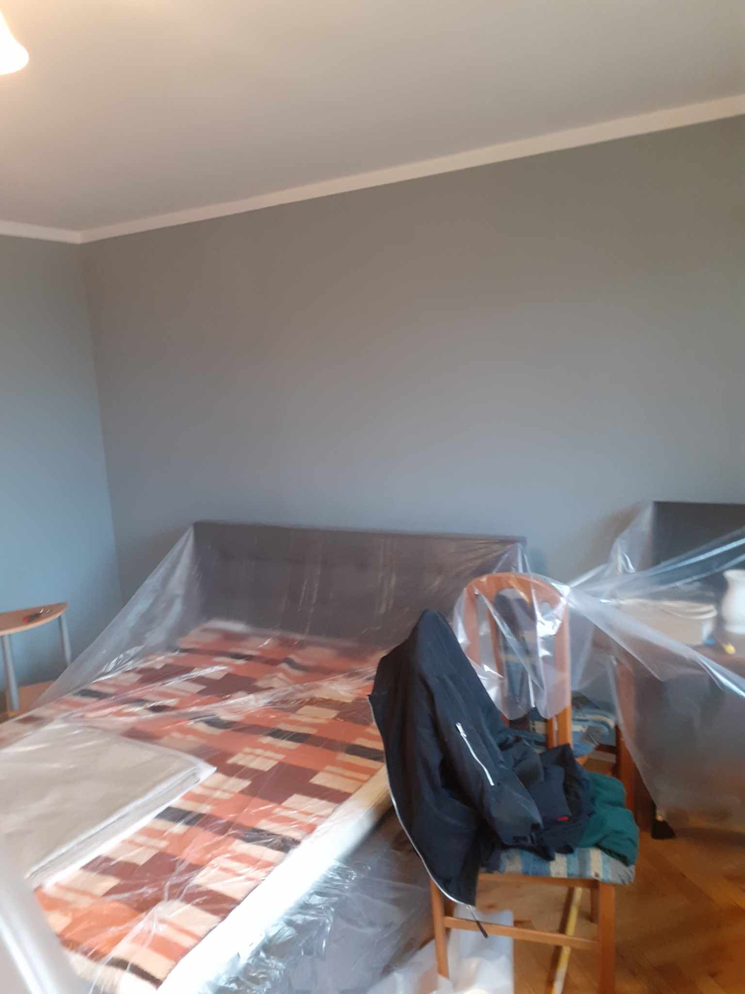 Pokój przed malowaniem: meble zabezpieczone folią, ściany w odcieniach szarości, listwa przysufitowa. Widok na przygotowane pomieszczenie do prac malarskich.