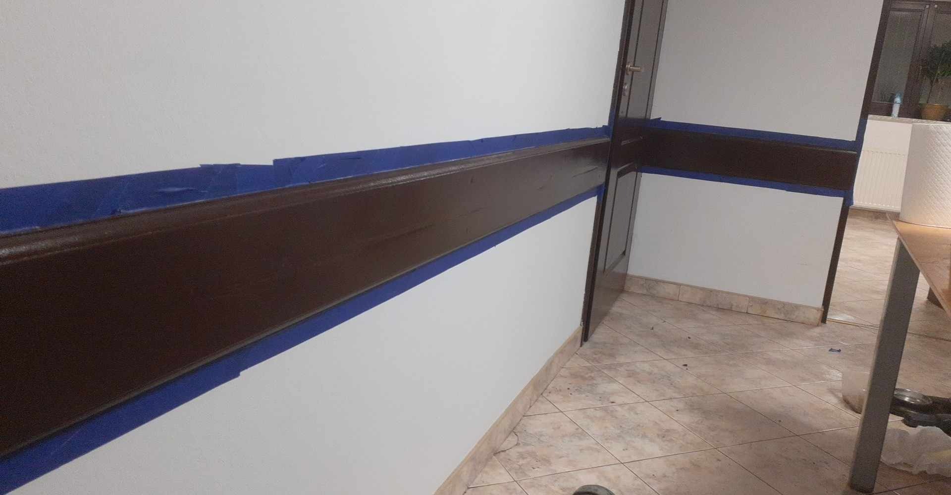 Świeżo pomalowana, brązowa lamperia zabezpieczona niebieską taśmą malarską na białej ścianie. Widoczna podłoga z płytek i fragment drzwi.