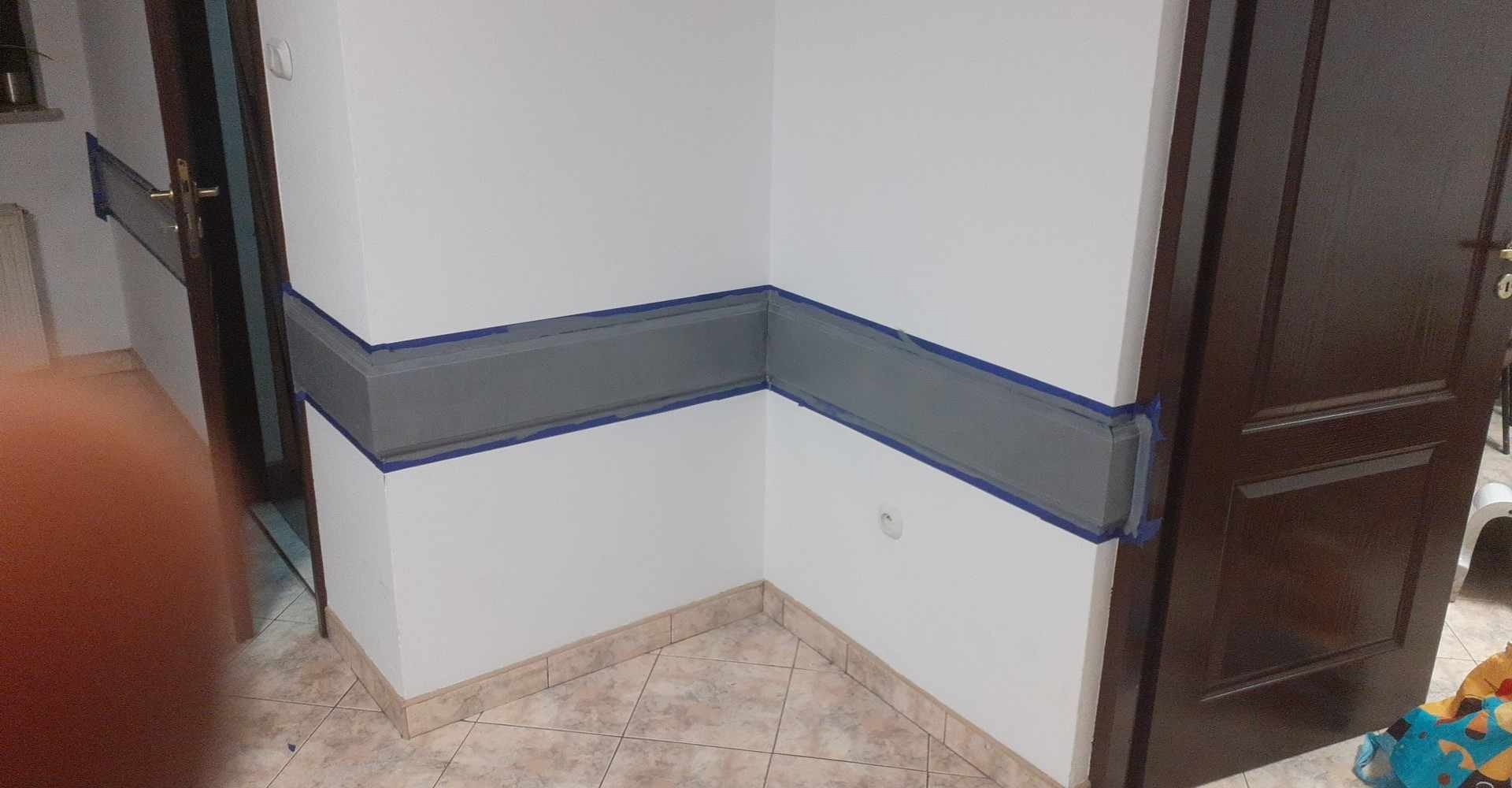 Przygotowanie do malowania: szara lamperia zabezpieczona taśmą malarską na białej ścianie w rogu pokoju. Widoczna podłoga z płytek i fragment otwartych, ciemnych drzwi.