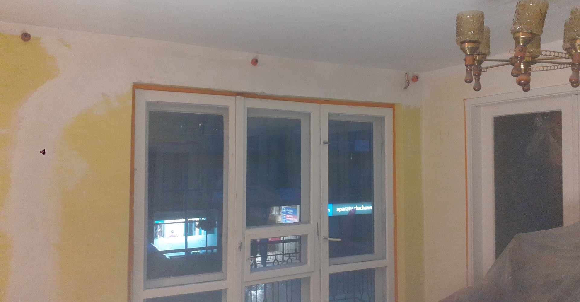 Ściana w trakcie malowania z widocznymi śladami szpachlowania, okno z białymi ramami i fragment żyrandola. Prace wykończeniowe w pomieszczeniu.