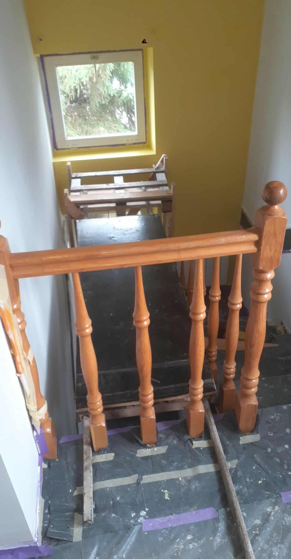 Malowanie klatki schodowej: pomalowane ściany na żółto, drewniana balustrada w trakcie malowania, zabezpieczona podłoga folią malarską. Widok z góry.