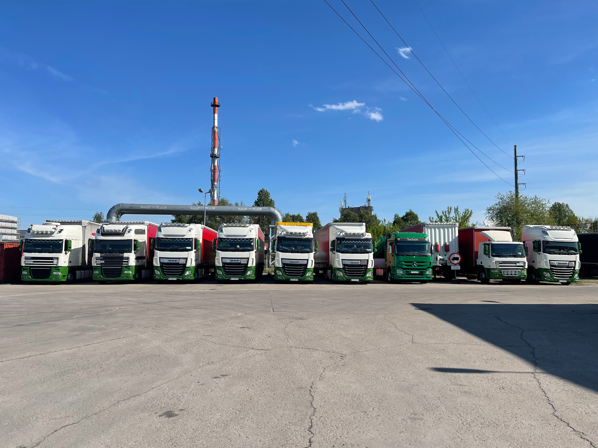 Flota ciężarówek DAF w barwach firmy, zaparkowana na placu z kominem w tle. Widoczne różne modele i zabudowy, w tym ciągniki siodłowe i pojedyncze ciężarówki.