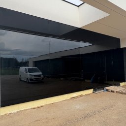 ELWENT DAWID KLIM - Elewacja budynku z czarnych paneli szklanych, odbijająca krajobraz i biały samochód dostawczy. Widoczne detale wykończeniowe i teren budowy.