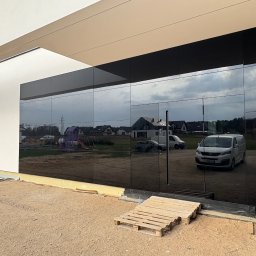 ELWENT DAWID KLIM - Elewacja budynku z czarnymi, lustrzanymi panelami odblaskowymi, ukazującymi odbicie otoczenia. Widoczne detale wykończeniowe i paleta budowlana.