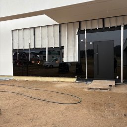 ELWENT DAWID KLIM - Montaż czarnej, szklanej fasady na nowoczesnym budynku. Widoczne aluminiowe profile i warstwa izolacyjna. Na ziemi piasek i akcesoria montażowe.