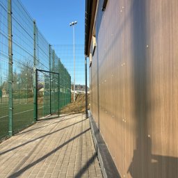 ELWENT DAWID KLIM - Nowoczesna elewacja budynku z drewnopodobnych paneli, widoczna z perspektywy chodnika obok boiska sportowego z zieloną siatką ogrodzeniową.