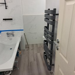 ELWENT DAWID KLIM - Wykończona łazienka z szarym grzejnikiem drabinkowym, białą wanną zabezpieczoną folią i marmurowymi płytkami na ścianach. Widoczne drewnopodobne panele podłogowe.