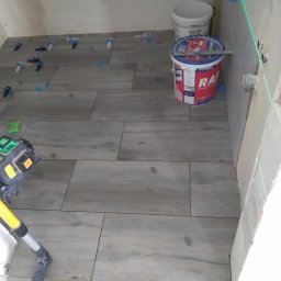 ELWENT DAWID KLIM - Układanie płytek podłogowych w łazience z użyciem poziomicy laserowej i systemu poziomowania. Widoczne wiaderko z klejem i płytki imitujące drewno.