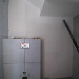 Szare płytki w łazience, widoczny stelaż podtynkowy WC, poziomica oparta o ścianę. Minimalistyczne wykończenie, surowy styl.