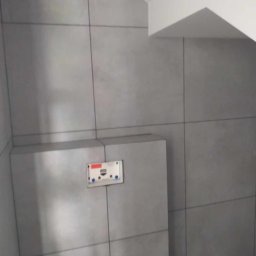 ELWENT DAWID KLIM - Wykończona łazienka z szarymi, matowymi płytkami wielkoformatowymi. Widoczny podtynkowy stelaż WC i fragment drewnopodobnej podłogi. Minimalistyczny design.