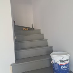 ELWENT DAWID KLIM - Pomalowane na szaro schody wewnątrz budynku. Na podeście wiadro z farbą. Widoczne narzędzia malarskie na schodach. Świeżo pomalowane ściany.