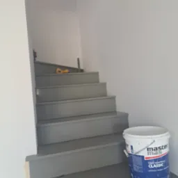 Pomalowane na szaro schody wewnątrz budynku. Na podeście wiadro z farbą. Widoczne narzędzia malarskie na schodach. Świeżo pomalowane ściany.