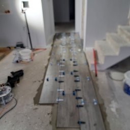 ELWENT DAWID KLIM - Układanie podłogi z płytek w korytarzu, z użyciem poziomujących klipsów. Widoczne narzędzia glazurnicze i schody w tle.