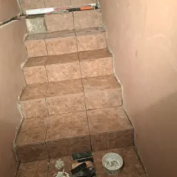 Wyłożone brązowe płytki na schodach wewnątrz budynku. Widoczne narzędzia glazurnicze i akcesoria do układania płytek. Częściowo wykończone schody.