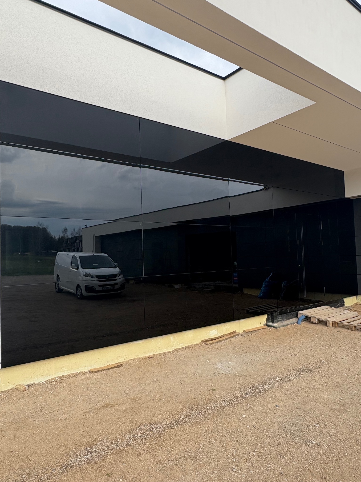 Elewacja budynku z czarnych paneli szklanych, odbijająca krajobraz i biały samochód dostawczy. Widoczne detale wykończeniowe i teren budowy.