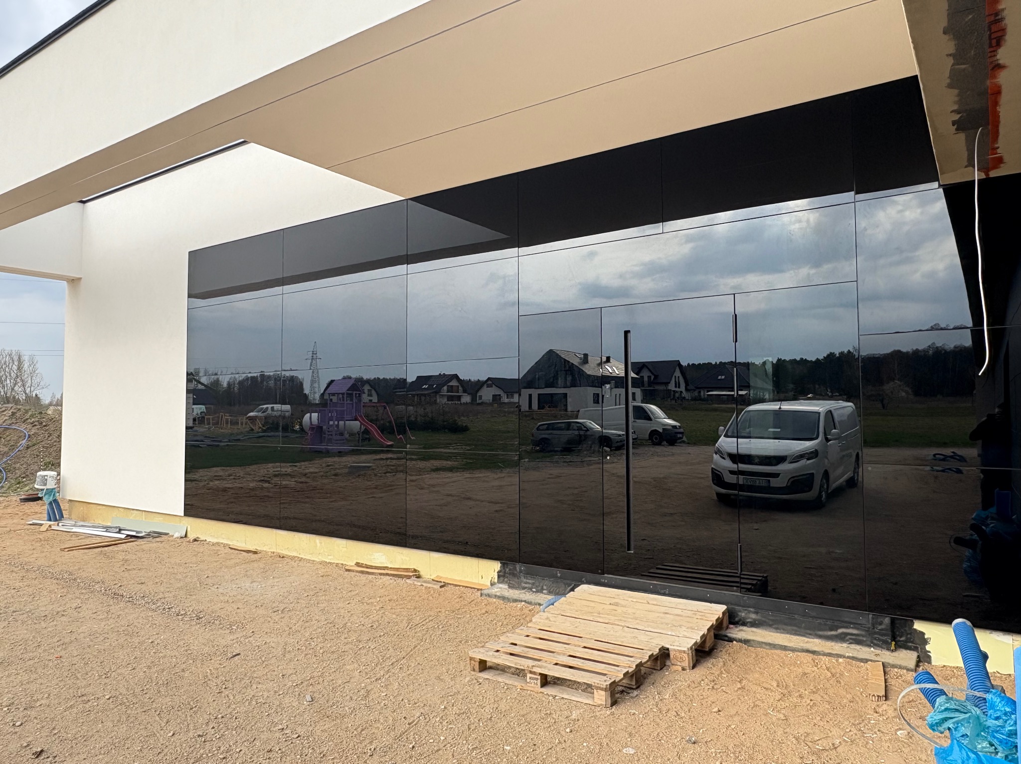 Elewacja budynku z czarnymi, lustrzanymi panelami odblaskowymi, ukazującymi odbicie otoczenia. Widoczne detale wykończeniowe i paleta budowlana.