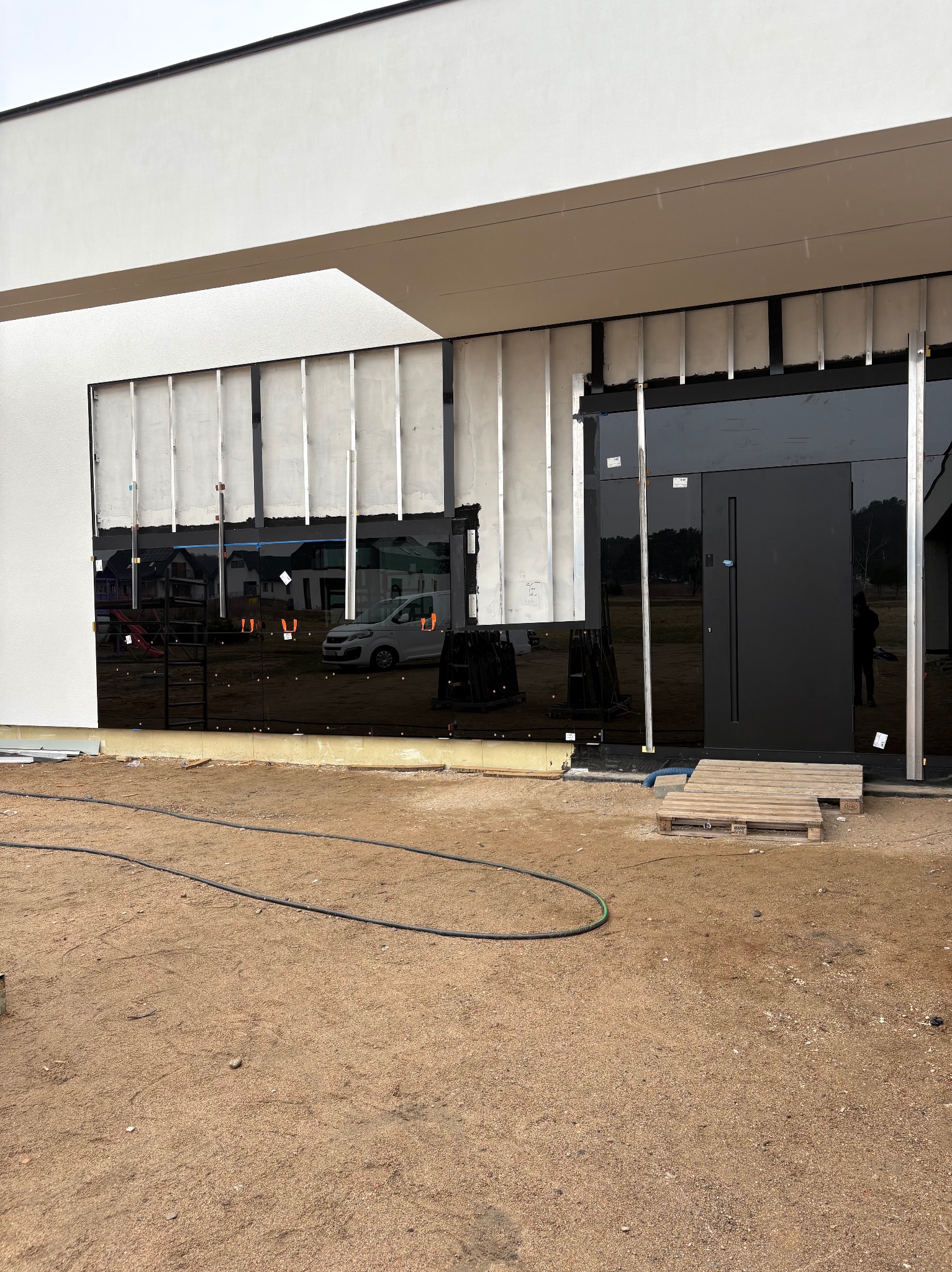 Montaż czarnej, szklanej fasady na nowoczesnym budynku. Widoczne aluminiowe profile i warstwa izolacyjna. Na ziemi piasek i akcesoria montażowe.