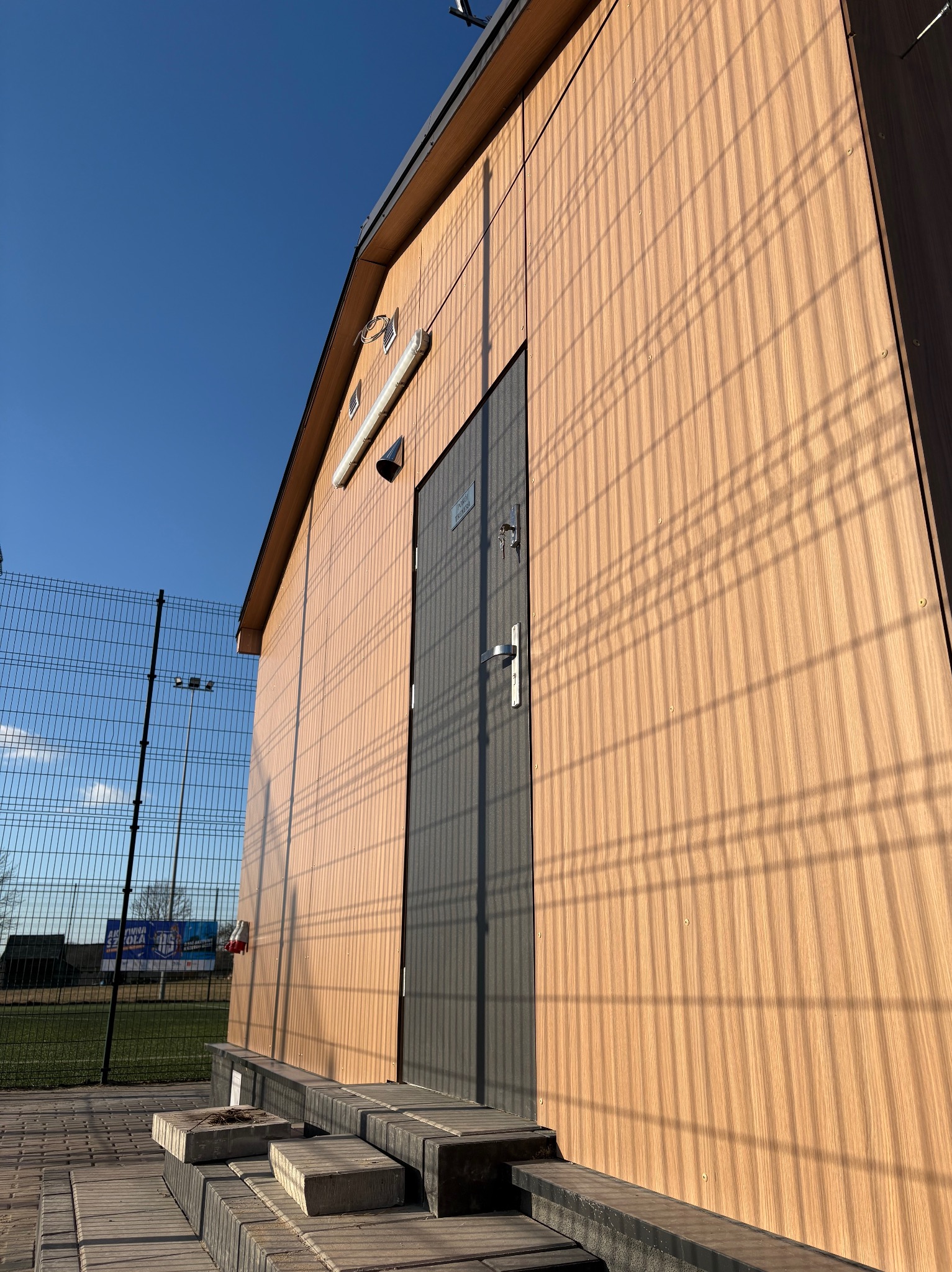 Elewacja budynku z paneli drewnopodobnych w ciepłym odcieniu, szara metalowa furtka wejściowa, widok z dołu, cień ogrodzenia na fasadzie, błękitne niebo.