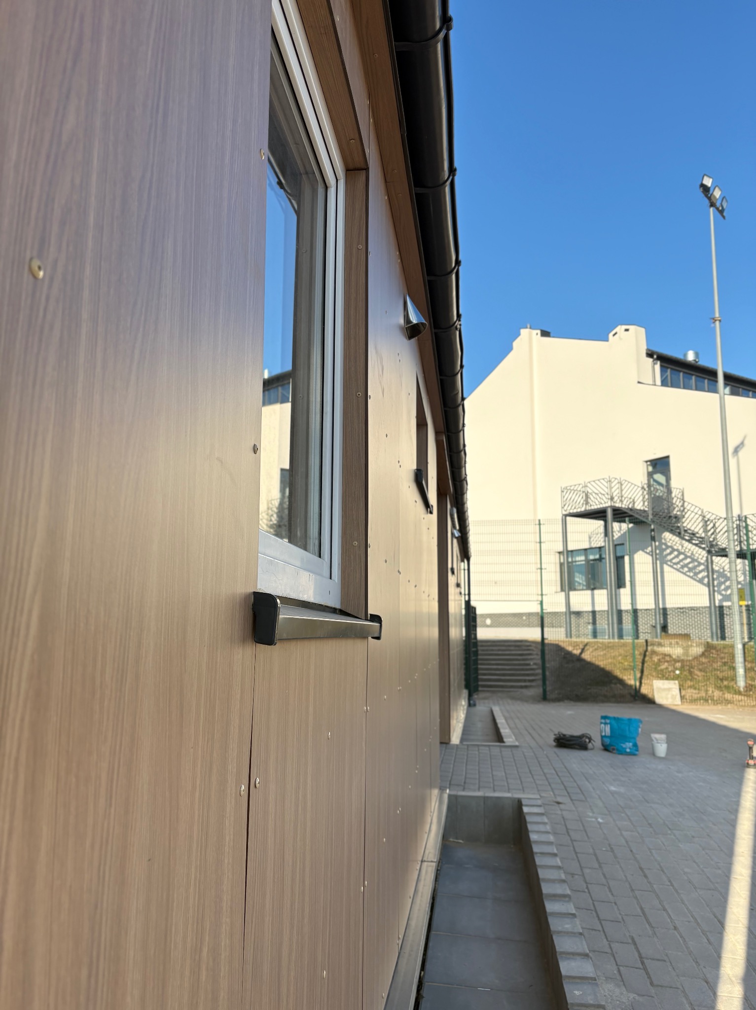 Wykończona elewacja budynku modułowego z oknem i orynnowaniem. Jasne, słoneczne tło z budynkiem i oświetleniem ulicznym. Widoczne detale łączeń paneli elewacyjnych.