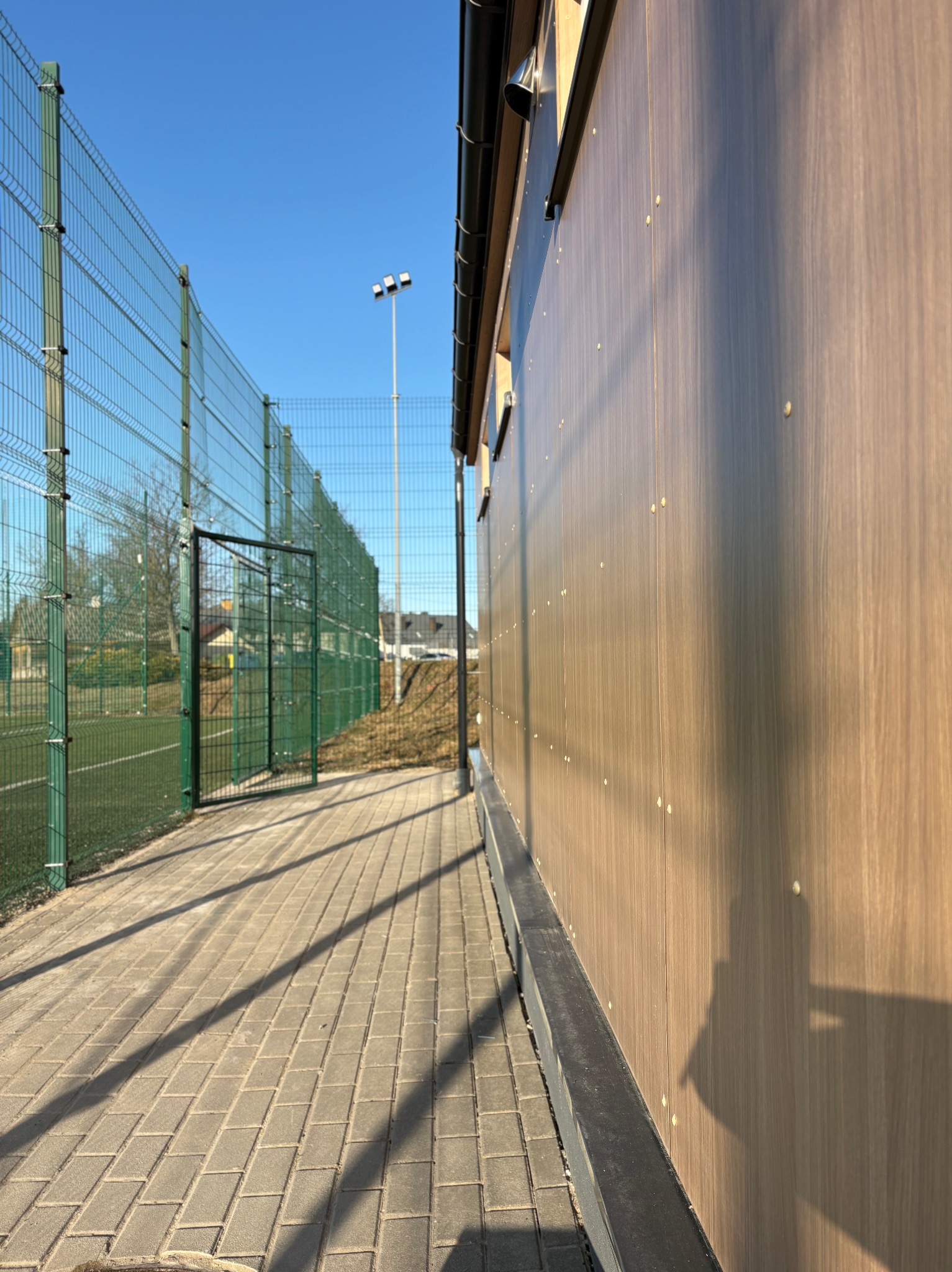 Nowoczesna elewacja budynku z drewnopodobnych paneli, widoczna z perspektywy chodnika obok boiska sportowego z zieloną siatką ogrodzeniową.