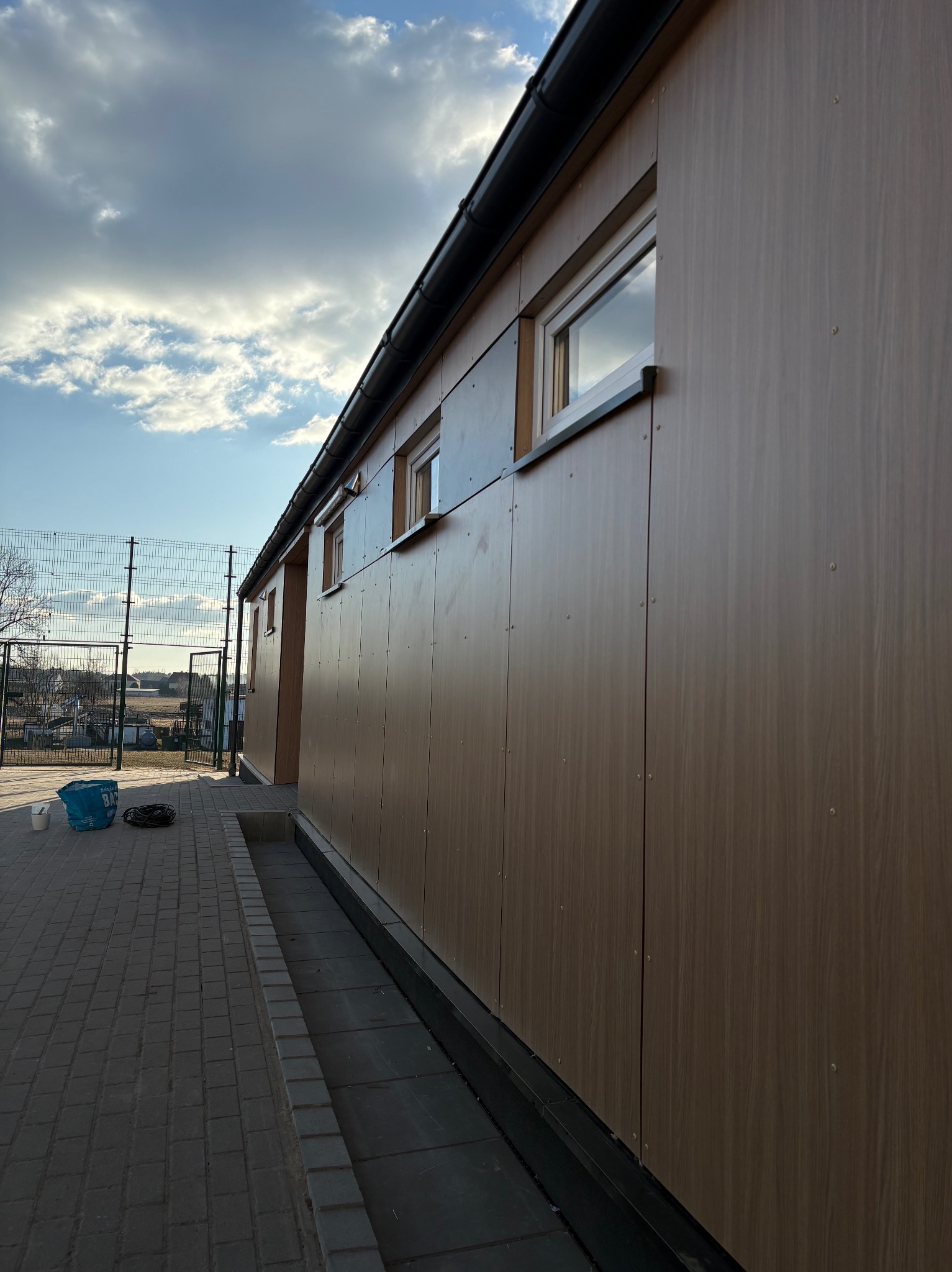 Elewacja budynku wykończona panelami imitującymi drewno, widoczne okna i elementy mocujące. W tle siatka ogrodzeniowa i błękitne niebo z obłokami.