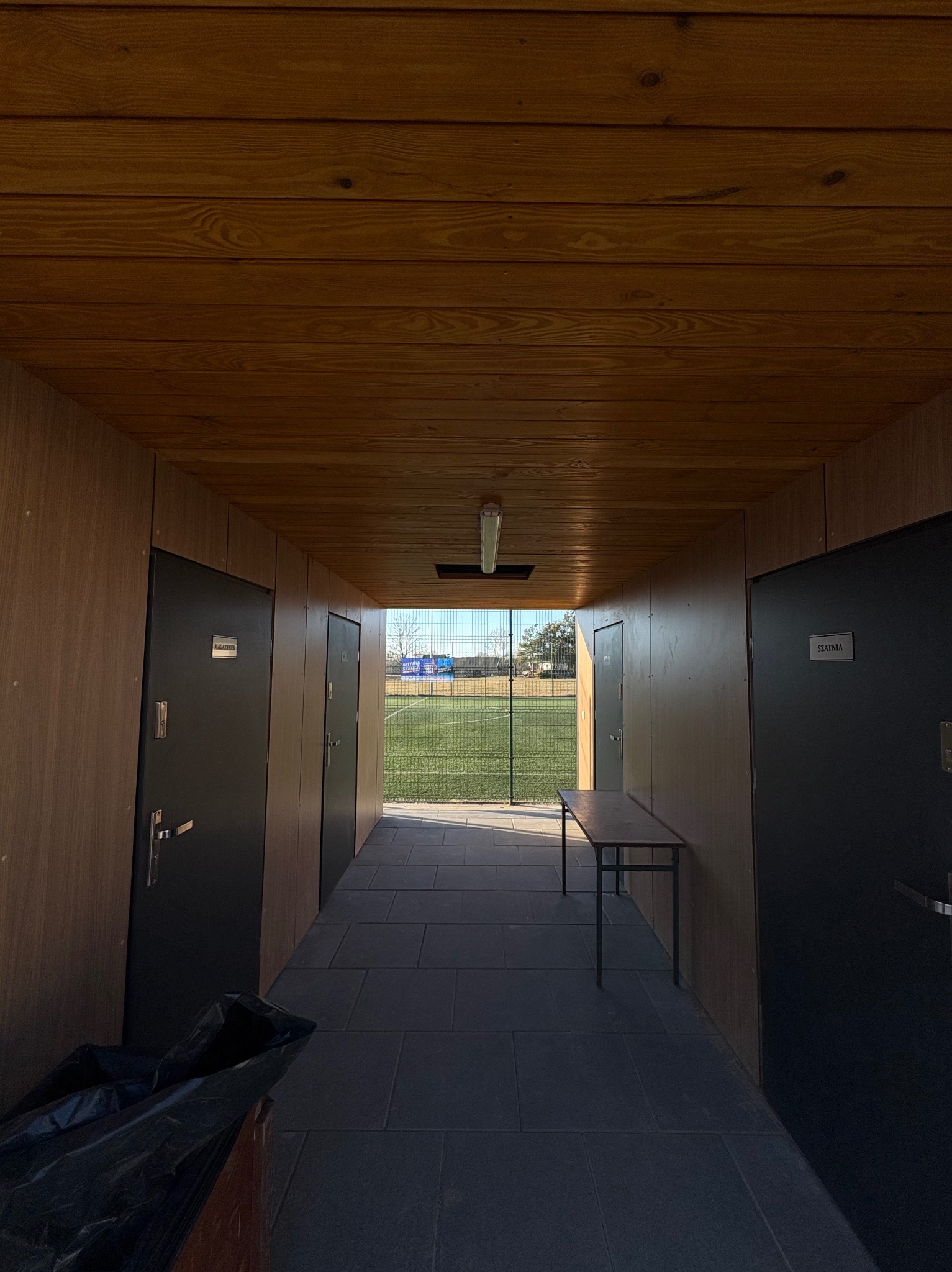 Korytarz z drewnianą boazerią i szatniami, widok na zewnątrz przez kratę. W tle boisko sportowe. Perspektywa od dołu, z widocznym stołem i workiem na śmieci.