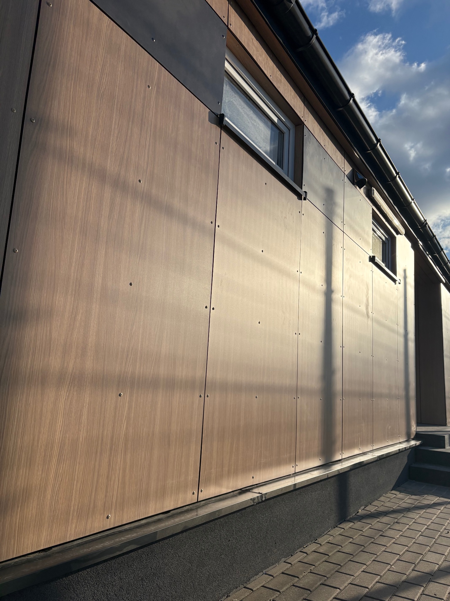 Fasada budynku wykończona panelami drewnopodobnymi z widocznymi mocowaniami. U góry okno i rynna. W tle błękitne niebo z chmurami.