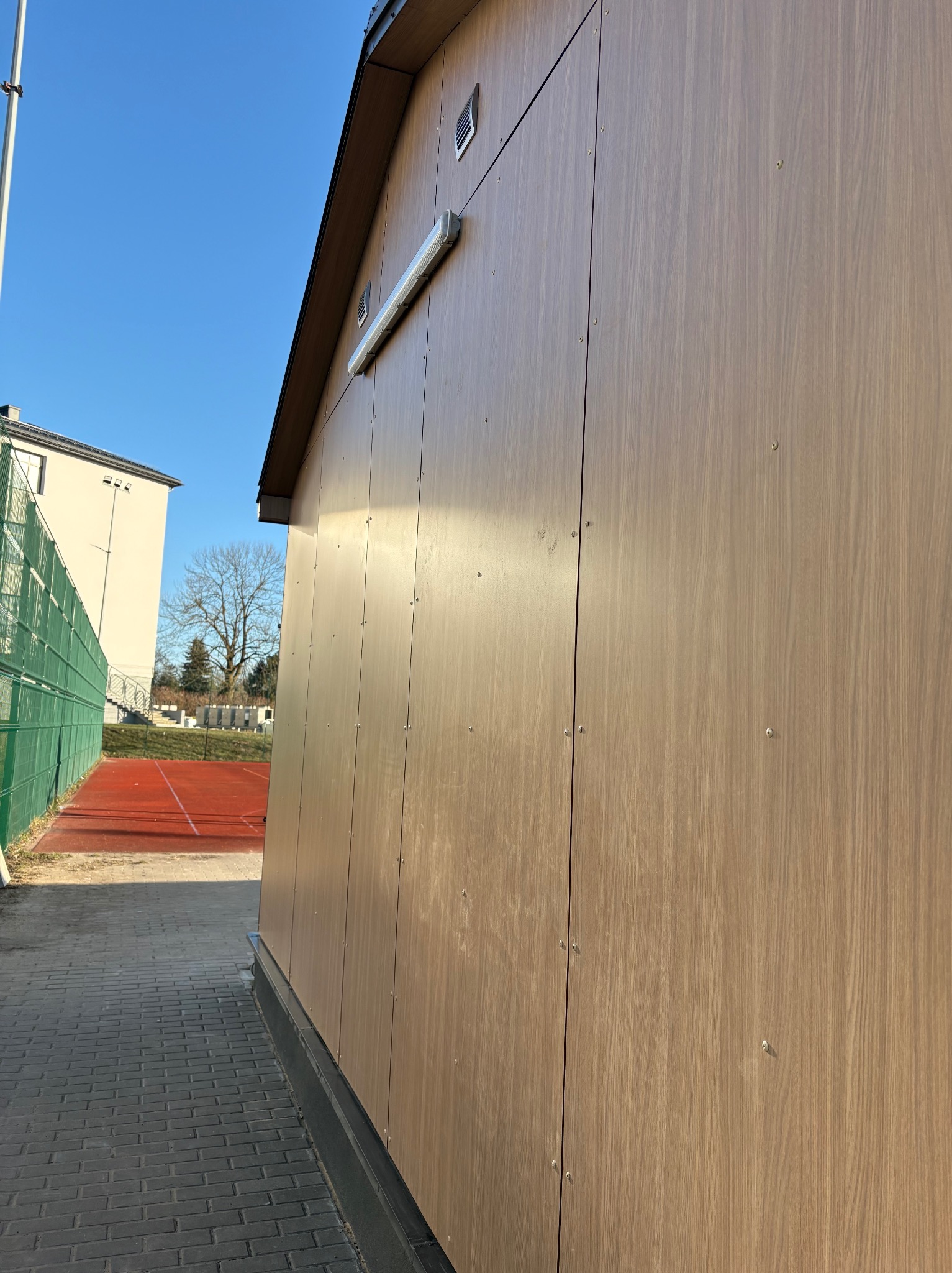 Wykończona elewacja budynku z paneli drewnopodobnych, widok z boku. Na tle błękitne niebo i fragment boiska sportowego. Widoczne elementy oświetlenia zewnętrznego.