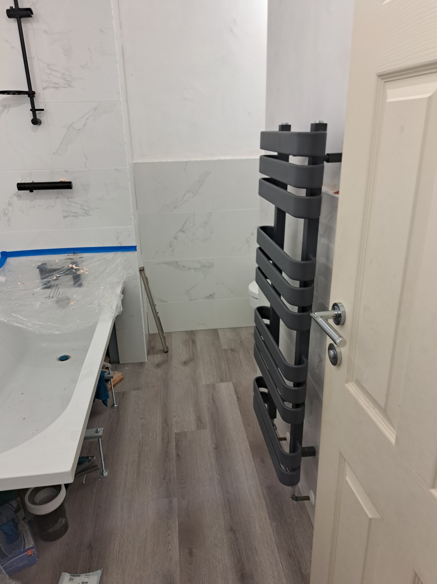 Wykończona łazienka z szarym grzejnikiem drabinkowym, białą wanną zabezpieczoną folią i marmurowymi płytkami na ścianach. Widoczne drewnopodobne panele podłogowe.