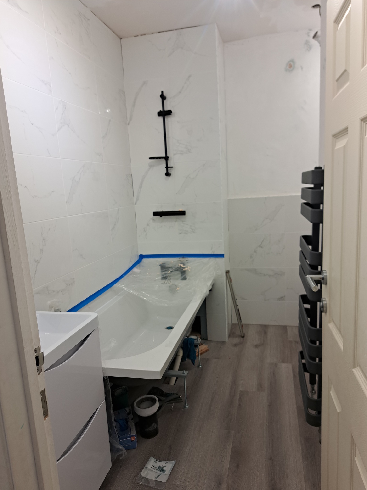 Biała łazienka z marmurowymi płytkami, nową wanną zabezpieczoną folią, grzejnikiem drabinkowym i szafką pod umywalkę, w trakcie lub tuż po remoncie.