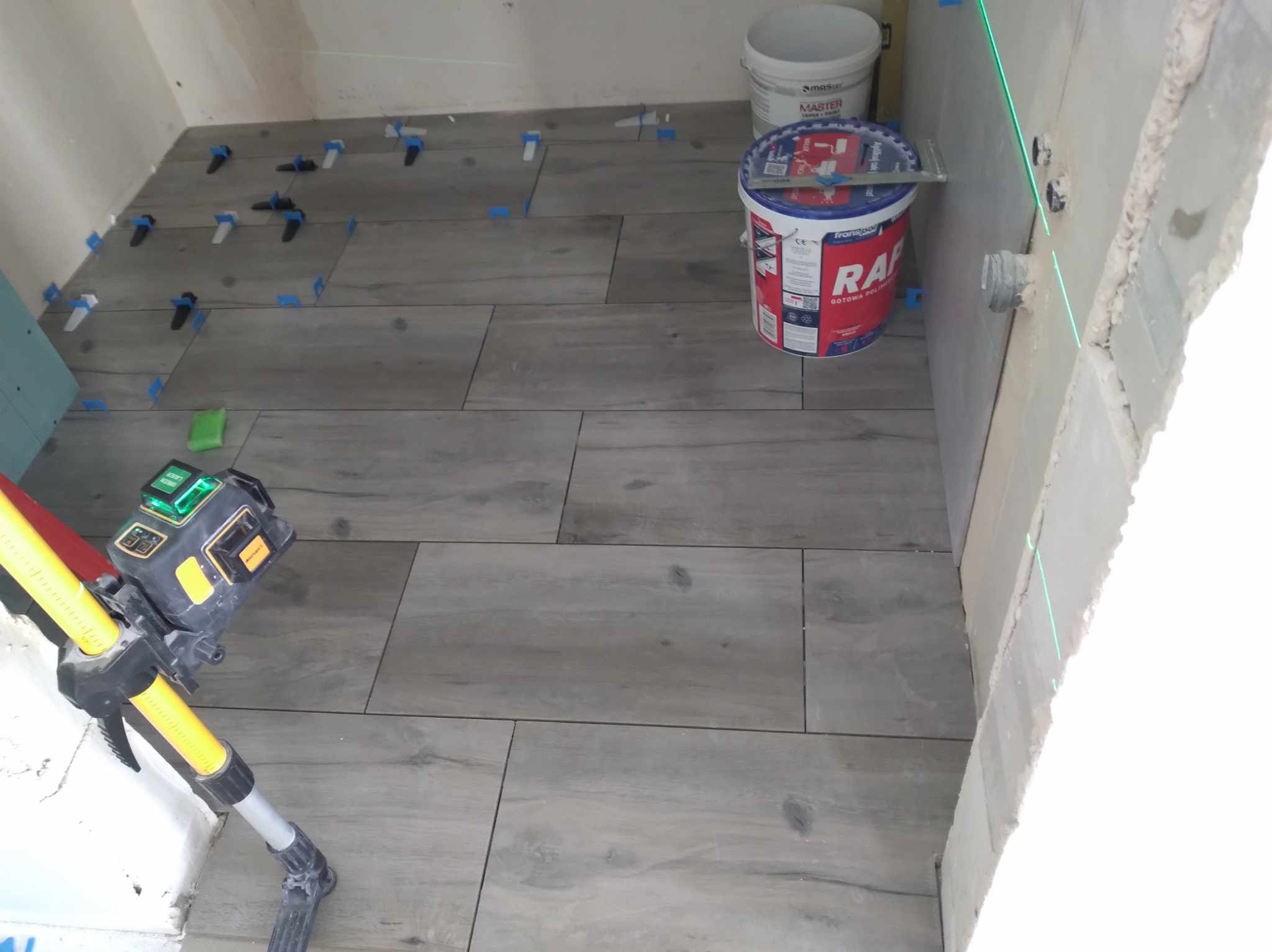 Układanie płytek podłogowych w łazience z użyciem poziomicy laserowej i systemu poziomowania. Widoczne wiaderko z klejem i płytki imitujące drewno.