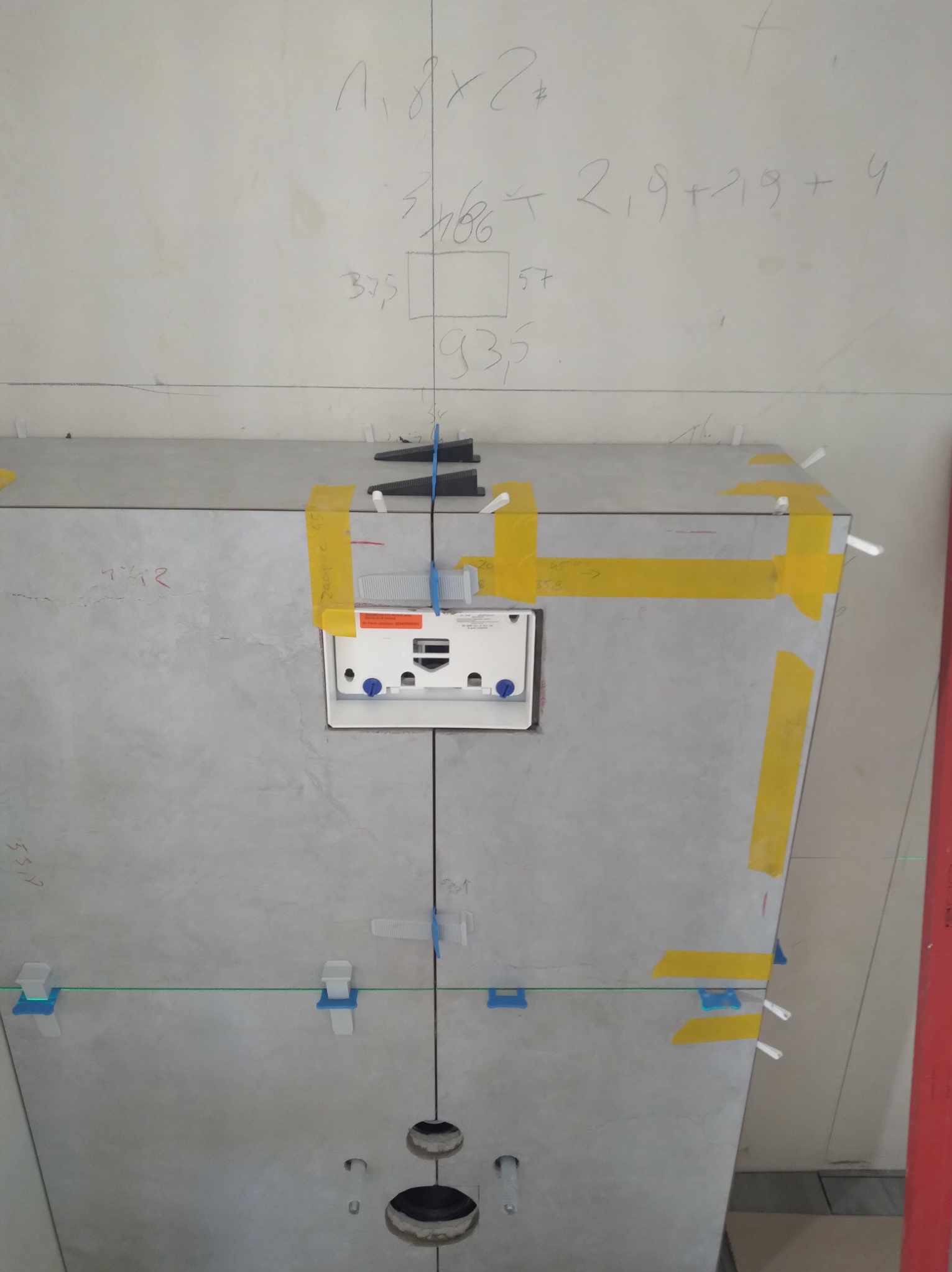 Szare płytki ścienne w trakcie montażu w łazience, z widocznymi elementami poziomowania i taśmą. Na ścianie notatki z wymiarami.