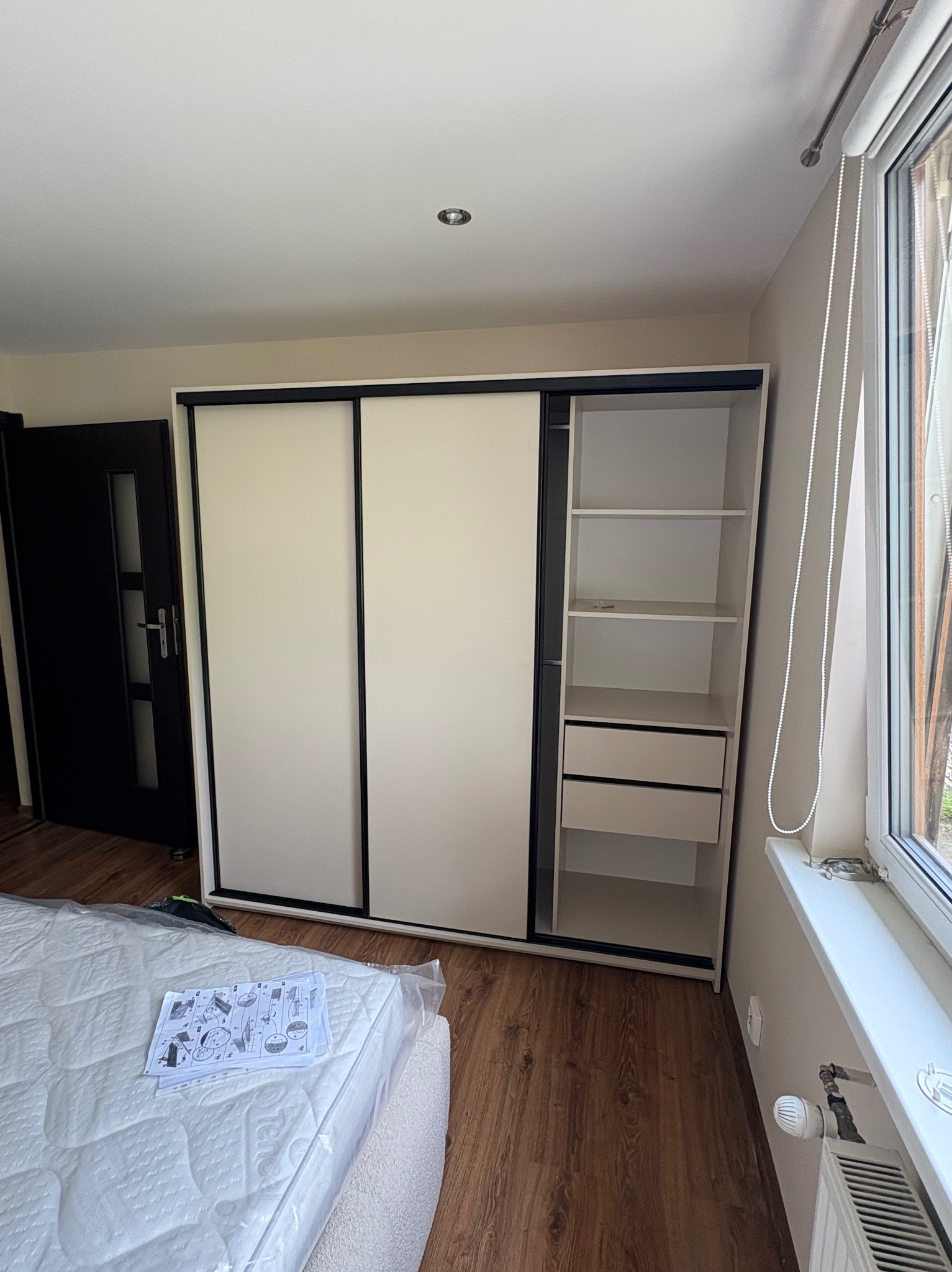 Nowoczesna szafa przesuwna z białymi frontami i czarnymi obramowaniami, częściowo otwarta, z widocznymi półkami i szufladami, na tle drewnianej podłogi.