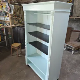 Błękitny regał z ciemnymi półkami stoi w warsztacie, widoczne narzędzia i materiały, co sugeruje ręczną produkcję mebla. Charakterystyczne toczone nogi.