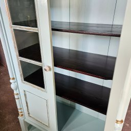 Reno-Art Warsztat Dawid Gąsiorek - Otwarta, odnowiona witryna w pastelowym odcieniu z ciemnymi półkami i przeszklonymi drzwiczkami. Widoczne złote akcenty i ceramiczny uchwyt.