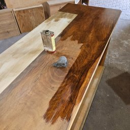 Reno-Art Warsztat Dawid Gąsiorek - Lakierowanie drewnianego blatu z widoczną puszką lakieru i wełną stalową. Część blatu surowa, część pokryta lakierem w odcieniach brązu. W tle elementy konstrukcji meblowej.