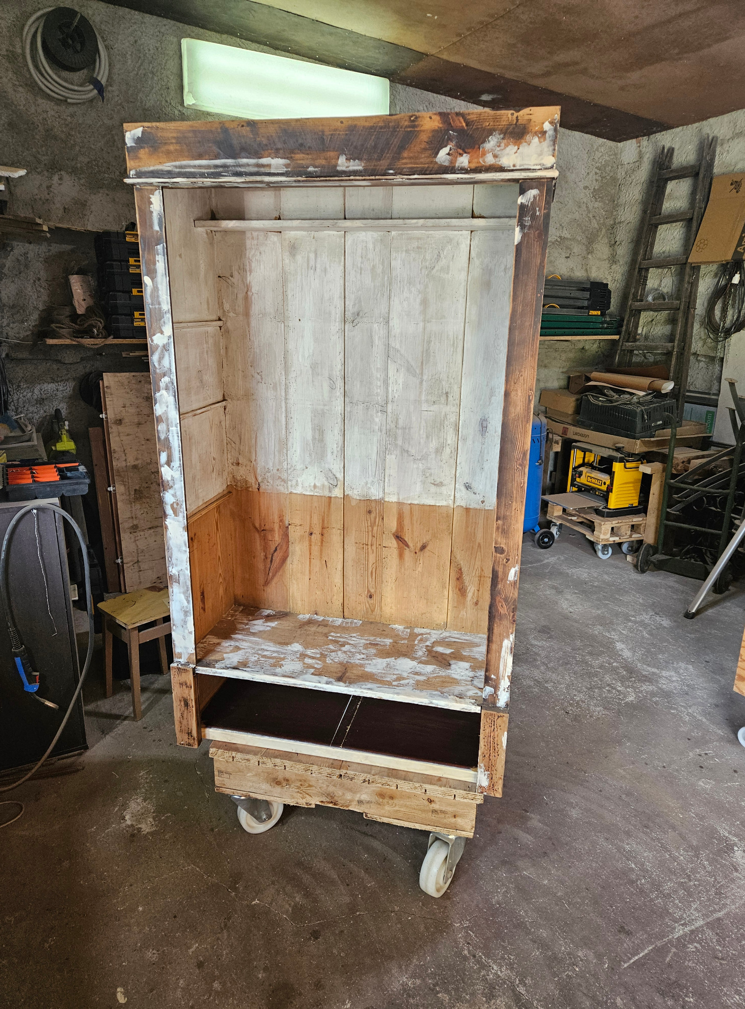 Renowacja szafy: drewniana konstrukcja na kółkach w trakcie odnawiania, widoczne ślady farby i szpachli, surowe tło warsztatu.