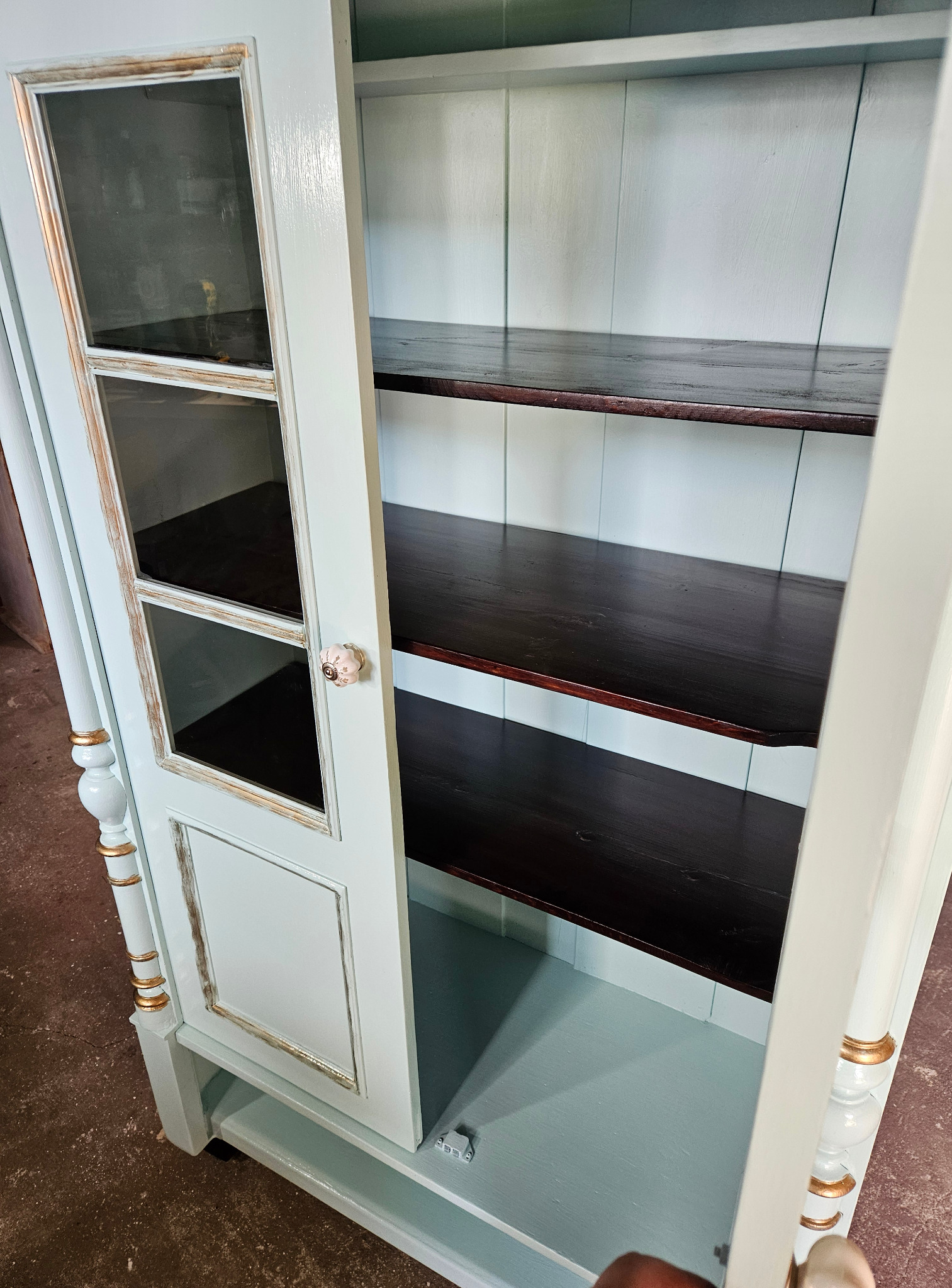 Otwarta, odnowiona witryna w pastelowym odcieniu z ciemnymi półkami i przeszklonymi drzwiczkami. Widoczne złote akcenty i ceramiczny uchwyt.