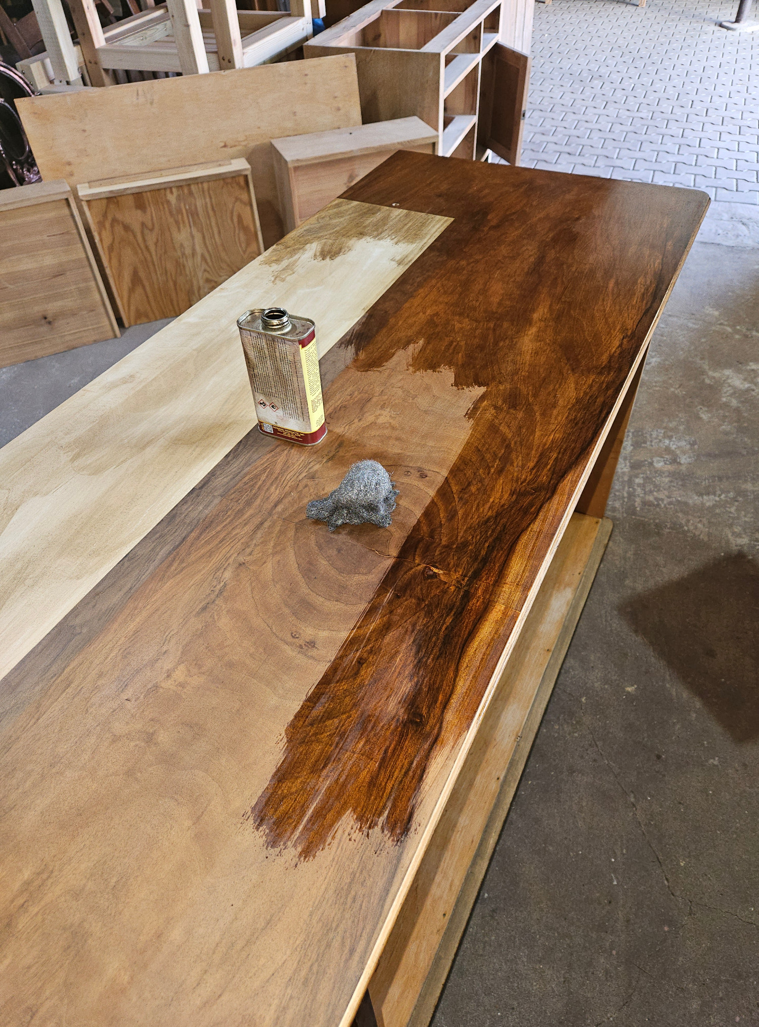 Lakierowanie drewnianego blatu z widoczną puszką lakieru i wełną stalową. Część blatu surowa, część pokryta lakierem w odcieniach brązu. W tle elementy konstrukcji meblowej.