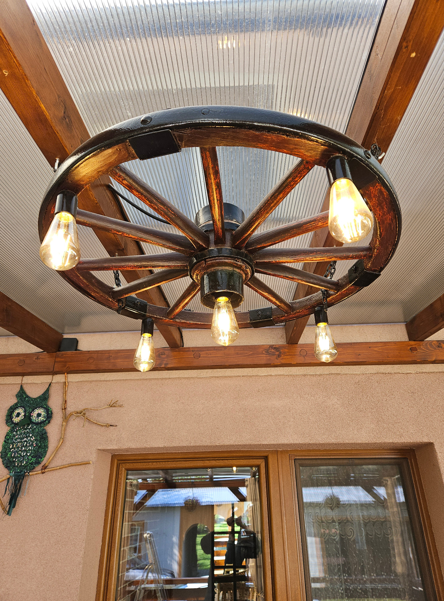 Stylowa lampa z koła od wozu, z żarówkami Edison, zamontowana na drewnianej konstrukcji w Środzie Wielkopolskiej. Dekoracyjny akcent z motywem sowy.