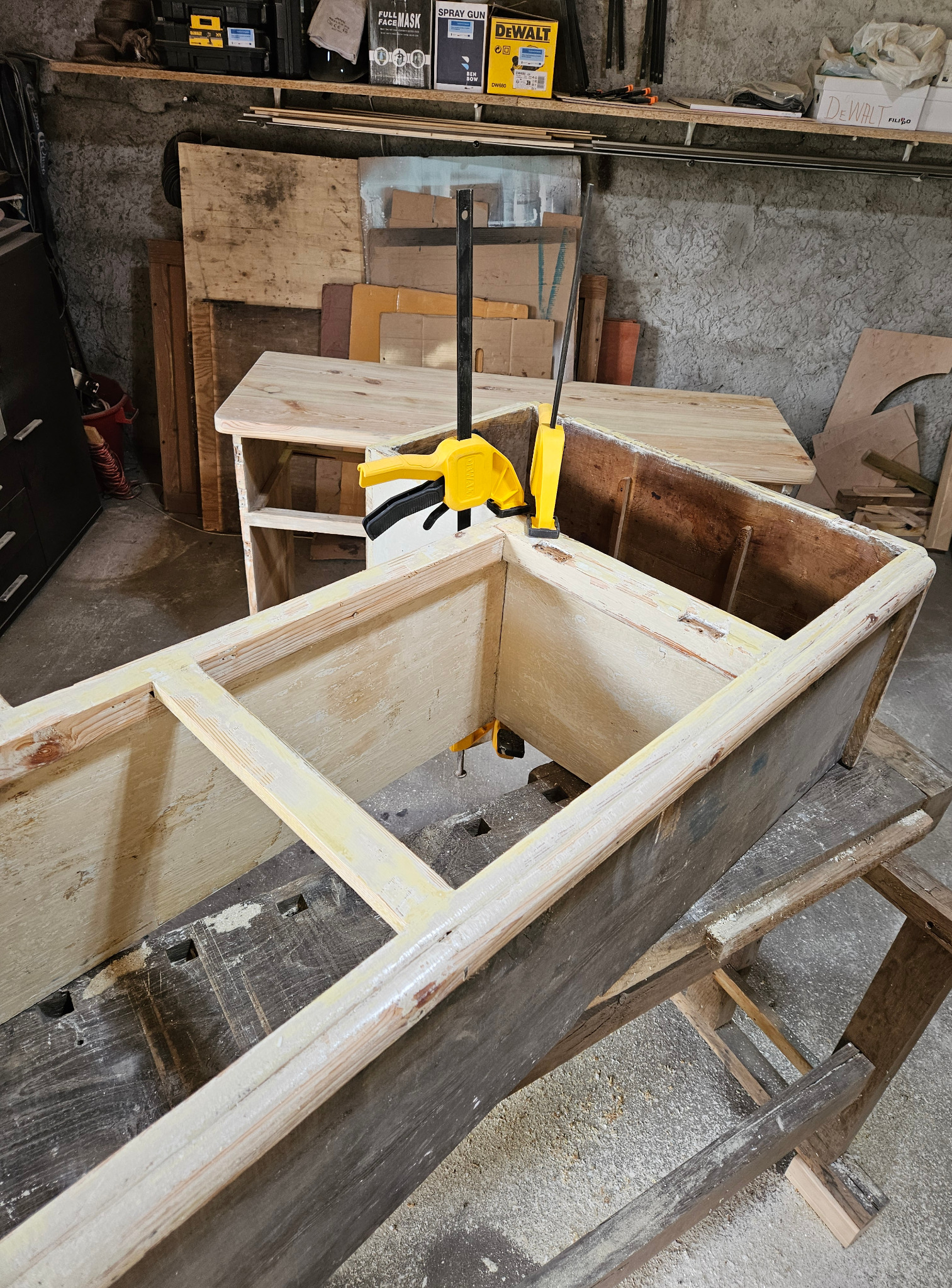 Rama drewnianej konstrukcji meblowej sklejona żółtym klejem i spięta ściskami stolarskimi, na tle warsztatu z drewnem i narzędziami.