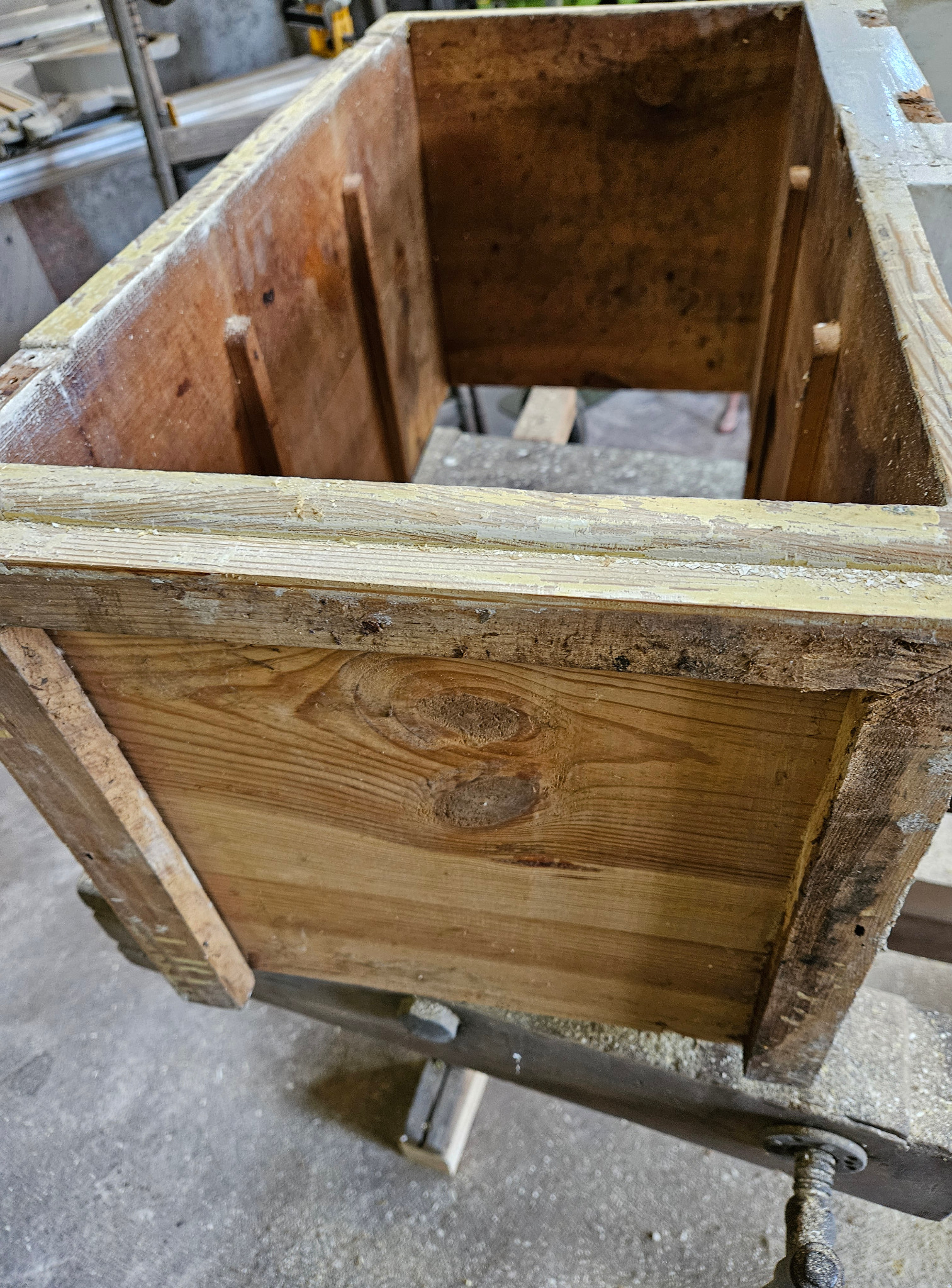 Drewniana skrzynia w warsztacie, mocowana w imadle, z widocznymi śladami użytkowania i resztkami farby, ukazująca proces renowacji lub naprawy mebla.