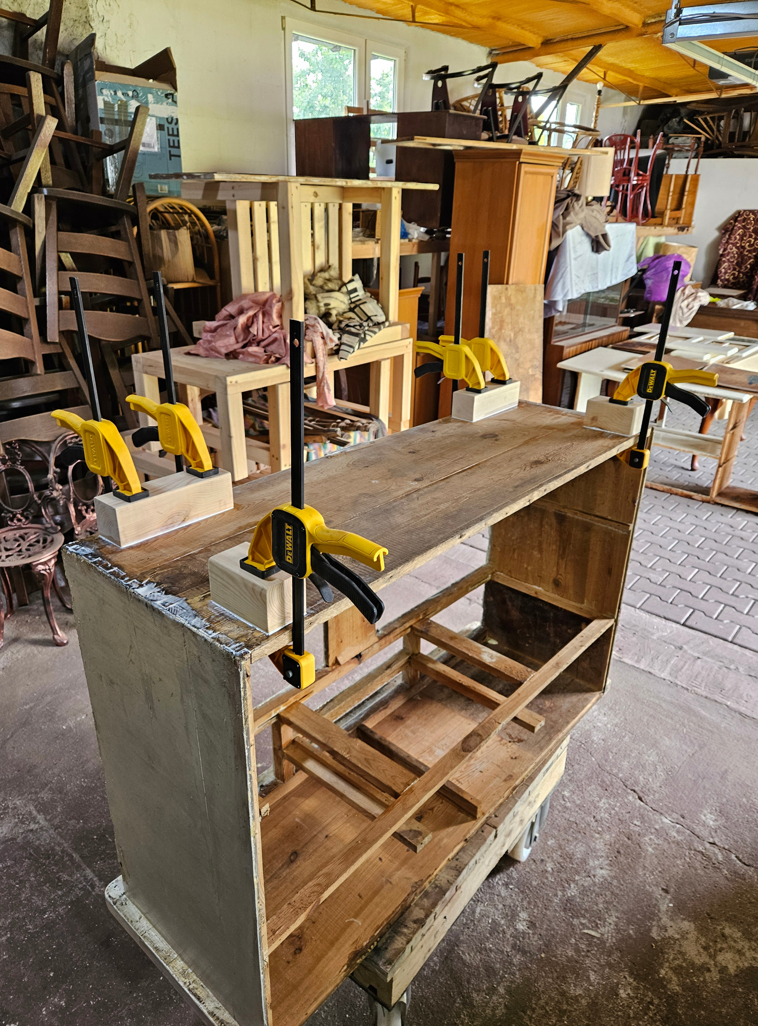 Renowacja drewnianej komody: żółte ściski stolarskie mocują elementy, widoczne ślady kleju. W tle warsztat z meblami i narzędziami, co sugeruje profesjonalne podejście.