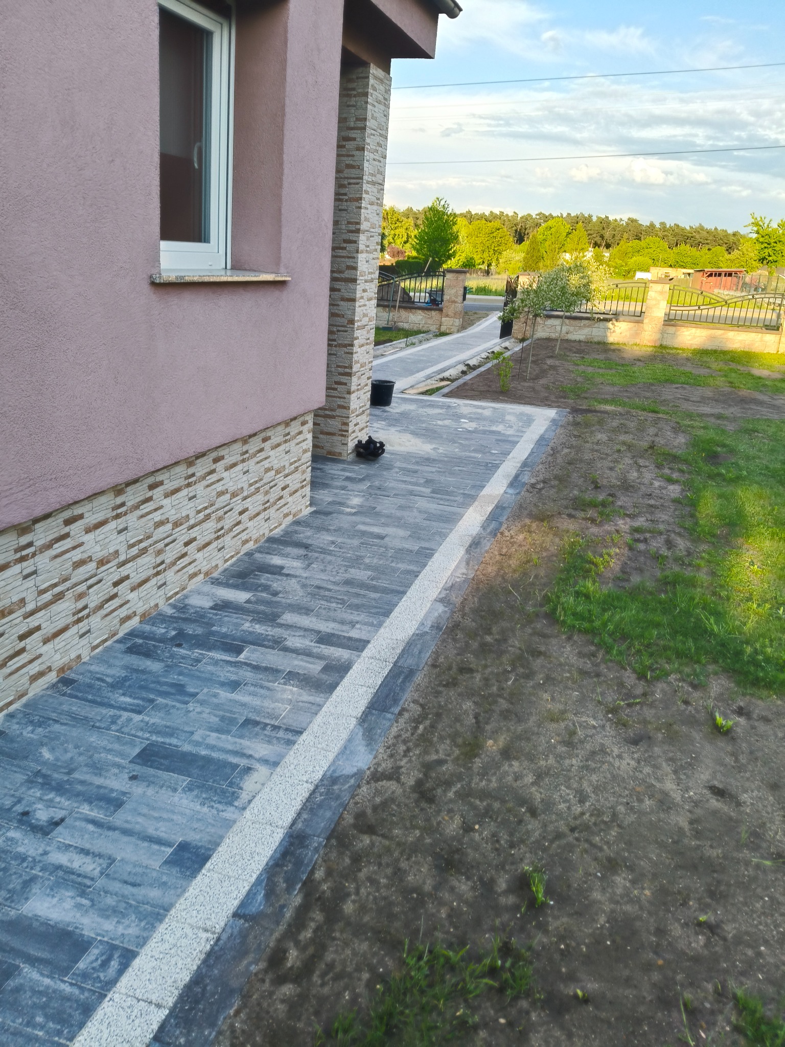 Ułożona, grafitowa kostka brukowa z jasnym obrzeżem przy budynku z elewacją w odcieniach beżu i różu. W tle widoczny trawnik i zieleń drzew.