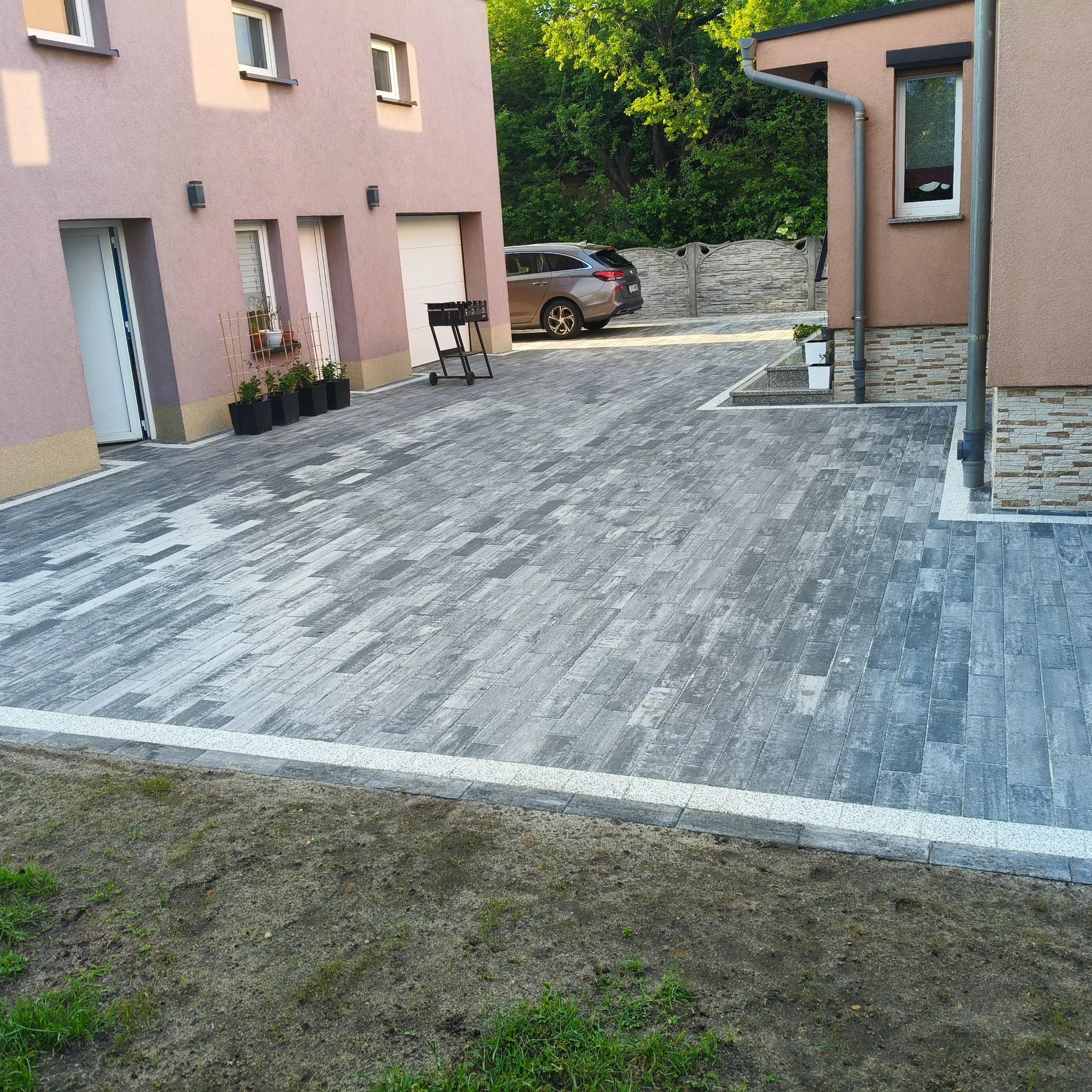 Ułożona szara kostka brukowa na podjeździe, obrzeża z jasnej kostki. W tle budynki z różową elewacją, samochód i grill. Widok z poziomu gruntu.