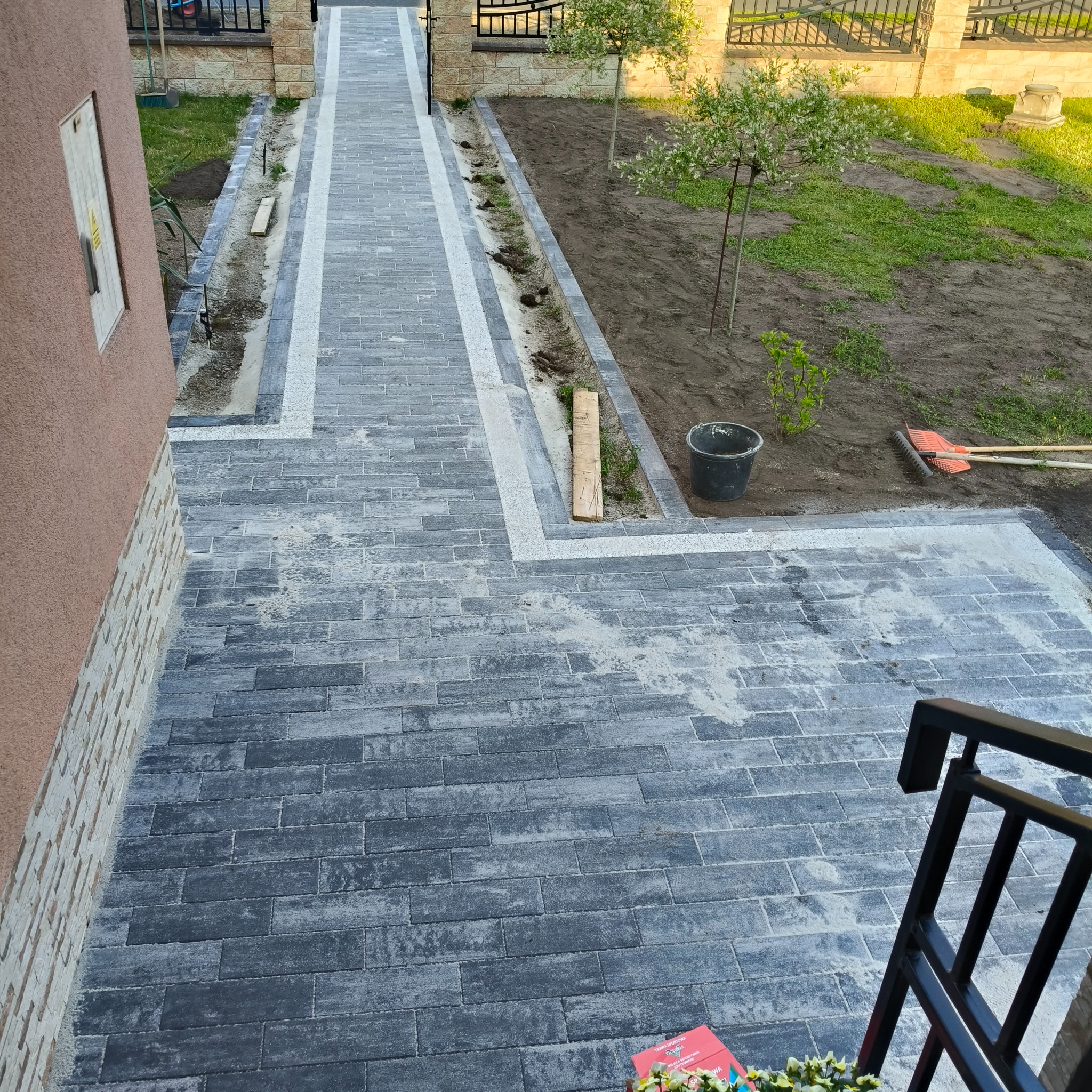 Ścieżka z szarej kostki brukowej z białymi obrzeżami, prowadząca wzdłuż budynku i rabatki z młodymi drzewkami. Widok z góry, z fragmentem balustrady.