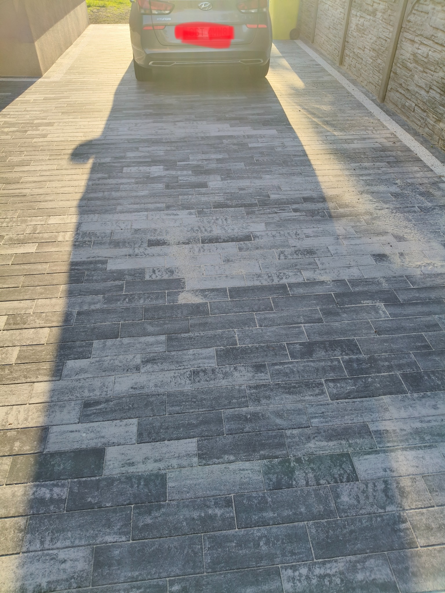 Ułożona szara kostka brukowa w kształcie prostokątów, cień samochodu i osoby, ogrodzenie z prawej strony. Widok z góry, w tle zaparkowany samochód.