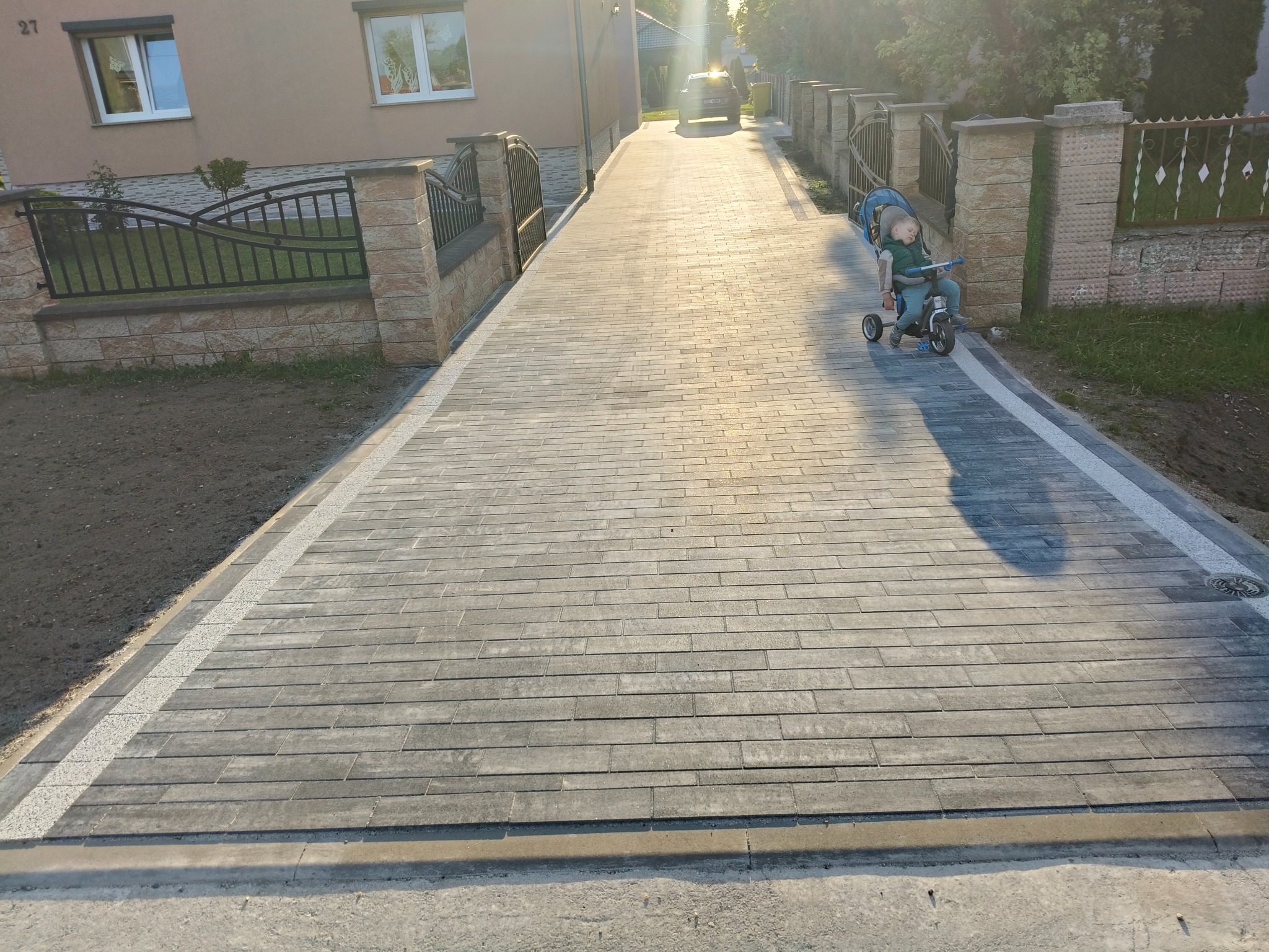 Długa, szara aleja z kostki brukowej prowadząca do domu z numerem 27. Po prawej stronie śpiące dziecko na trójkołowym rowerku. Jasne słońce na końcu drogi.