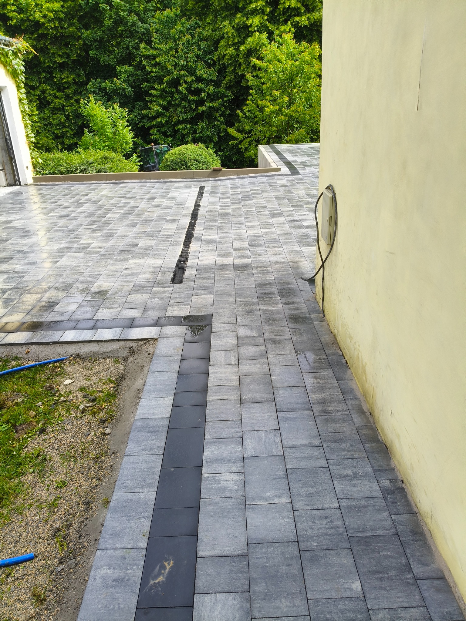 Nawierzchnia z szarej kostki brukowej ułożonej wzdłuż ściany budynku z elementami drenażu liniowego. W tle widoczna zieleń i fragment budynku.