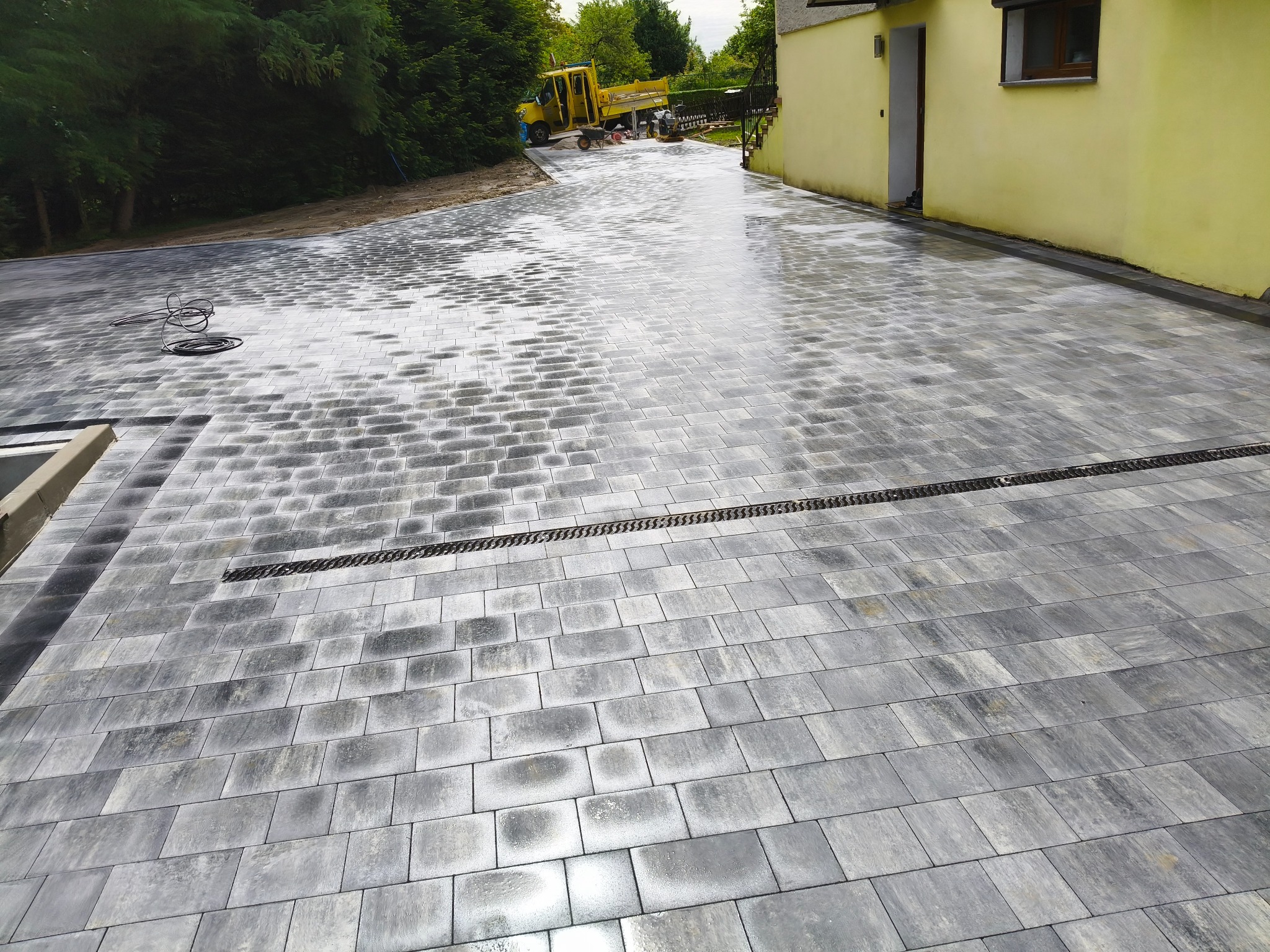 Ułożona, szara kostka brukowa z odpływem liniowym przed budynkiem z żółtą elewacją. Na dalszym planie zaparkowany żółty samochód dostawczy i zieleń.